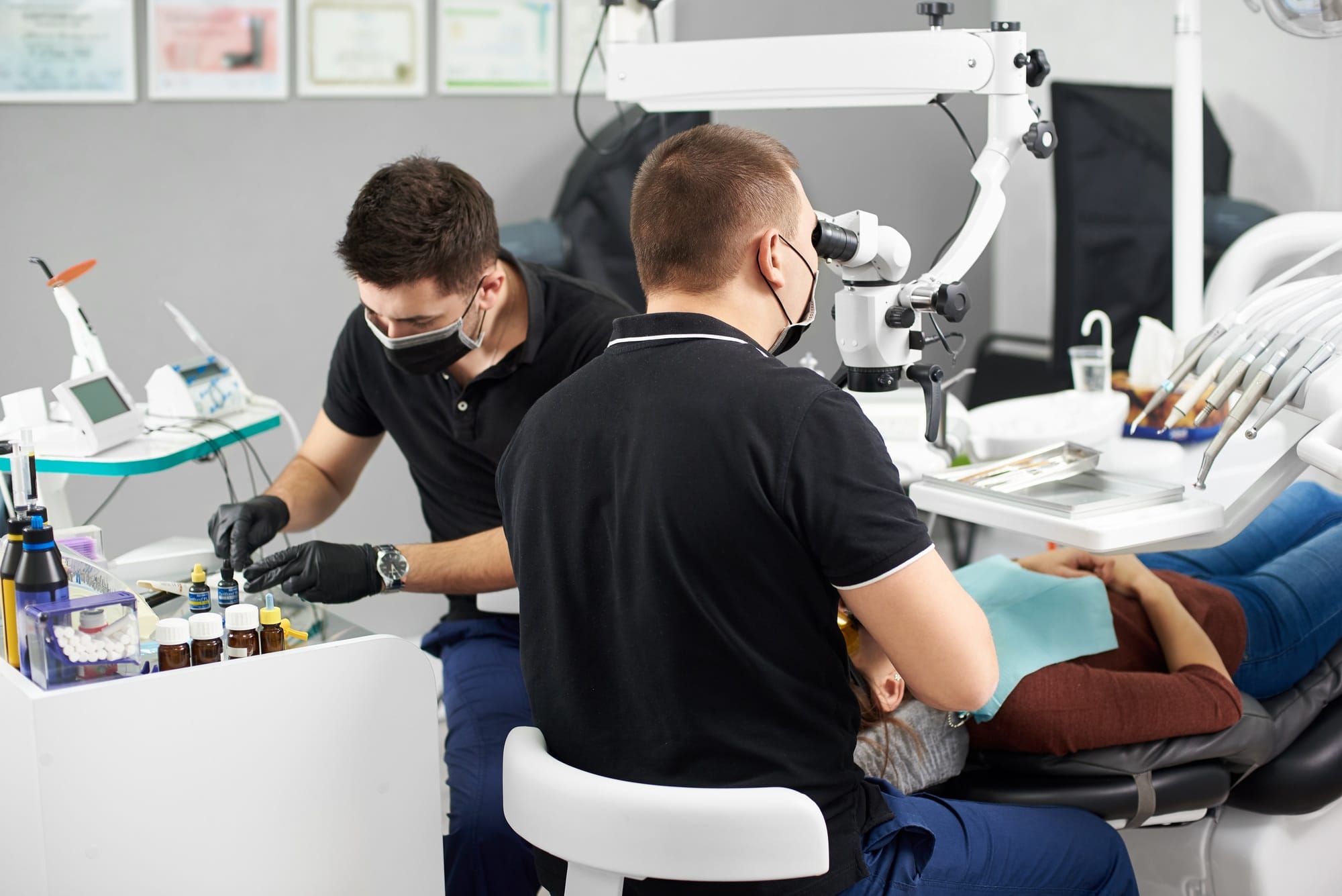 One male dentist is fixing a patient's teeth while another dentist is preparing the dental filling