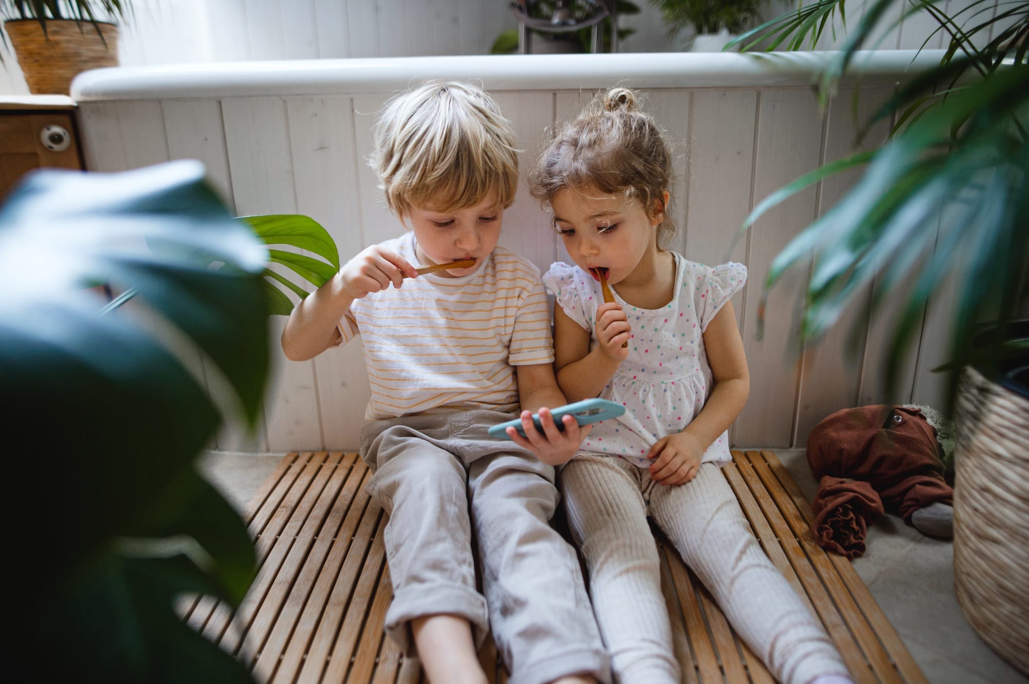 Cute small children sitting on floor indoors at bathroom, brushing teeth and using smartphone Cute small children sitting on floor indoors at bathroom, brushing teeth and using smartphone