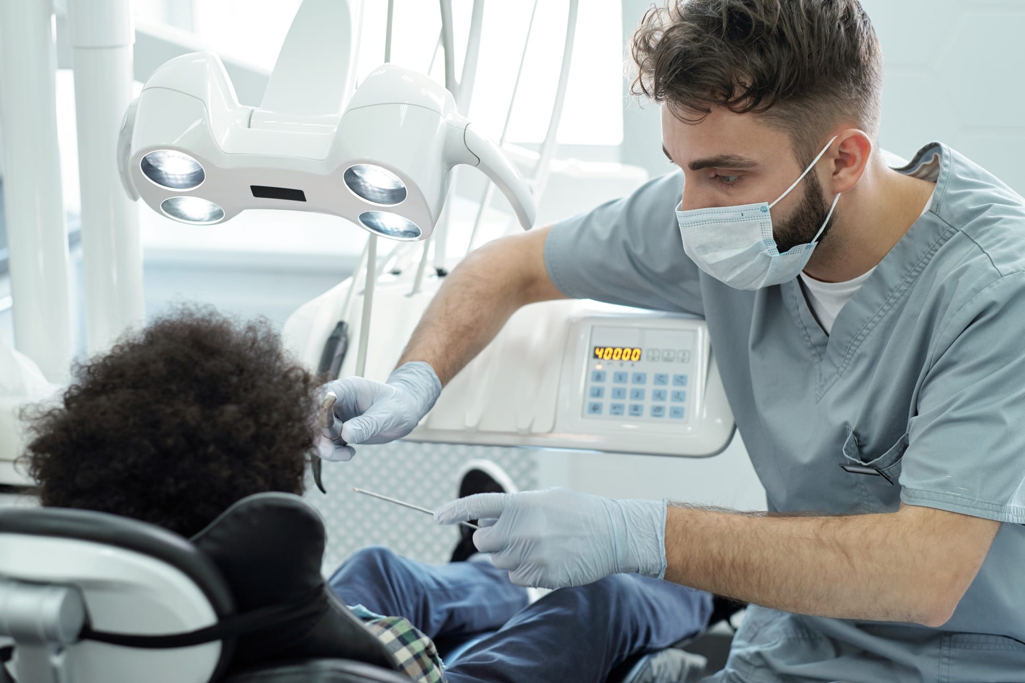 Dentist in uniform, mask and gloves holding dental drill and instrument by mouth of little patient Dentist in uniform, mask and gloves holding dental drill and instrument by mouth of little patient