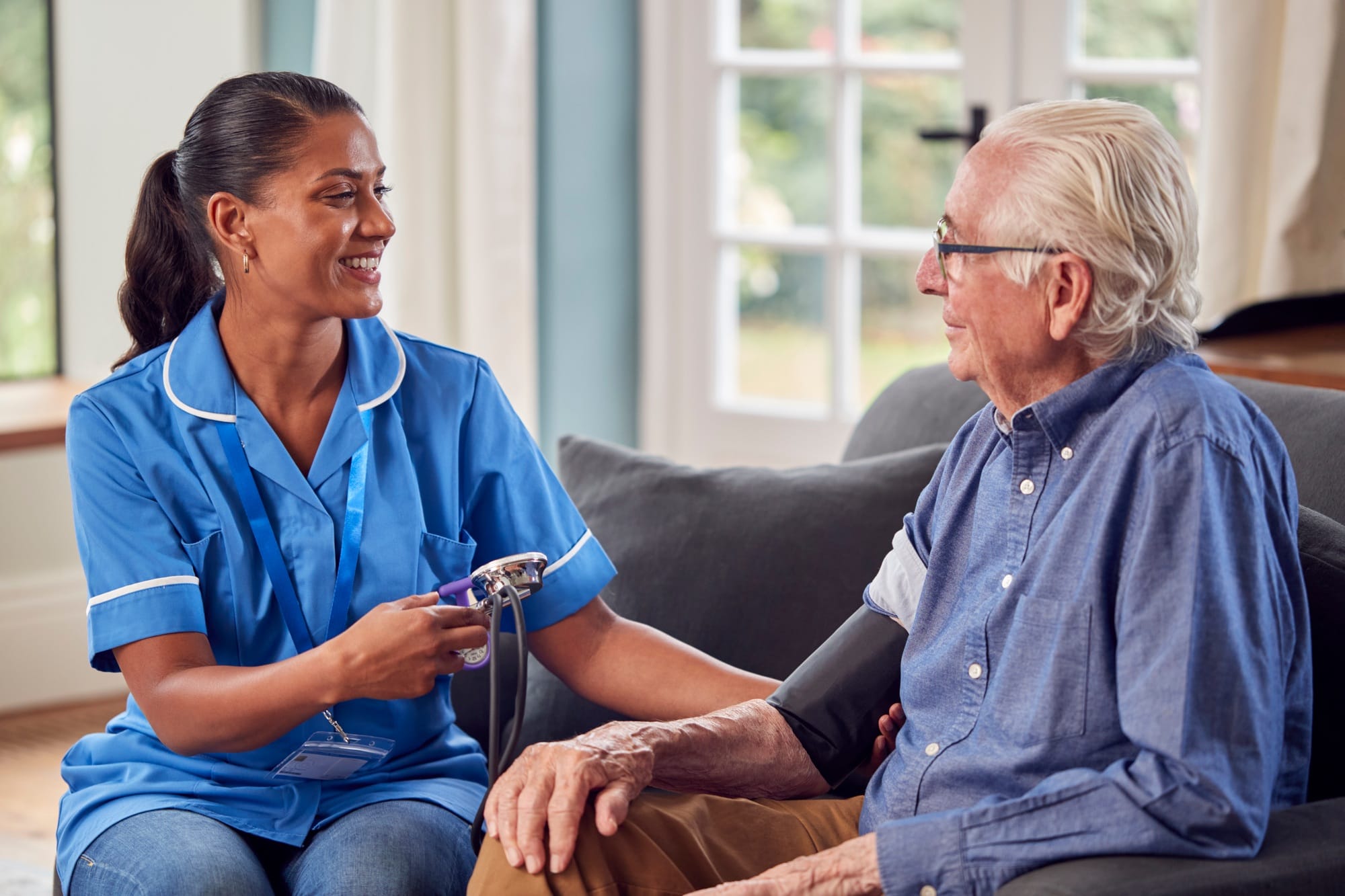 Senior Man At Home In Lounge Having Blood Pressure Taken By Female Care Worker In Uniform Senior Man At Home In Lounge Having Blood Pressure Taken By Female Care Worker In Uniform