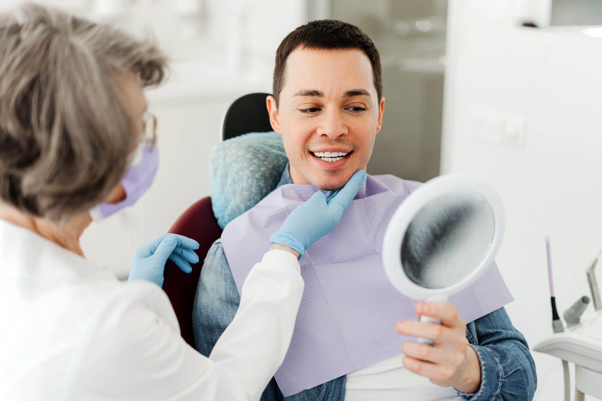 Smiling attractive male patient holding mirror, looking, talking to senior professional dentist Smiling attractive male patient holding mirror, looking, talking to senior professional dentist